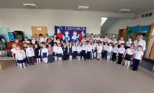 dzieci w strojach galowych z kotylionami przypiętymi lub zawieszonymi na szyi stoją w półkolu z nimi pani w tle dekoracja 11 listopada