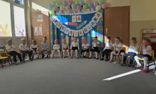 dzieci w eleganckich strojach stoją w półkolu w tle dekoracja z kredkami i balonami z napisem pasowanie na przedszkolaka
