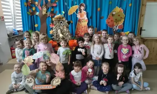 grupa dzieci pozuje do zdjęcia na tle jesiennej dekoracji wśród dzieci stoi figura jeża wykonana z papieru jeden z chłopcó trzyma napis dzień jeża