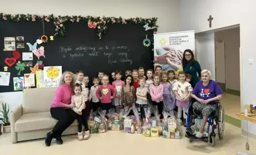 grupa dzieci wraz z paniami i panią na wózku inwalidzkim pozują do zdjęcia grupowego na tle wystroju przed dziećmi paczki z różnymi produktami spożywczymi