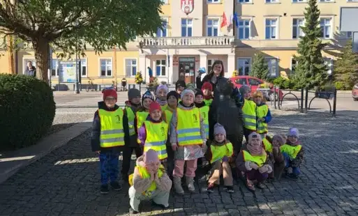 grupa dzieci w kamizelkach odblaskowych wraz z panią pozuje do zdjęcia z figurą pomnikiem jeża w tle widać Urząd Miasta Zgierza