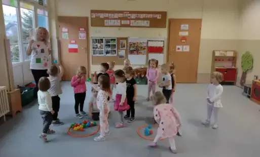 dzieci wraz z panią stoją na sali na podłodze leżą dwie obręcze w których są kolorowe piłeczki