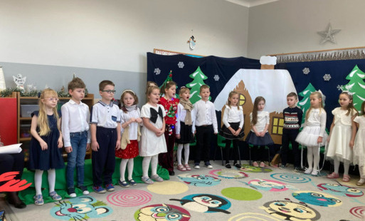 dzieci w odświętnych strojach stoją w półkolu za nimi świąteczno zimowa dekoracja