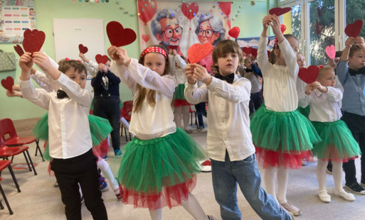 dzieci w odświętnych ubraniach stoją na sali unosząc w górę czerwone serca