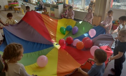 dzieci trzymają dookoła chustę animacyjną na której leżą kolorowe balony