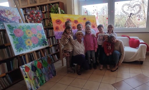 dzieci wraz z paniami pozują do zdjęcia kucając obok widać ustawione obrazy na których namalowane są kwiaty w tle dekoracje w oknie 