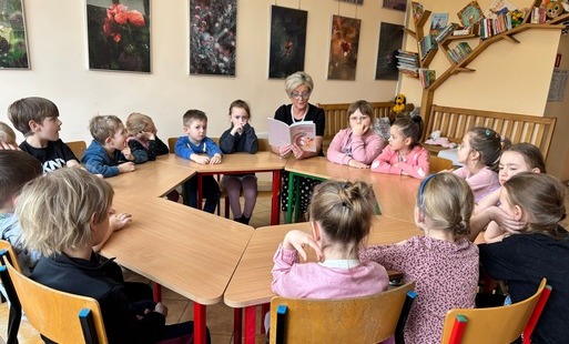 dzieci siedzą przy stolikach ustawionych w koło wśród nich siedzi pani trzymająca książkę