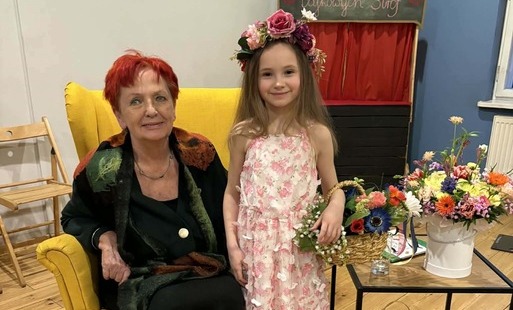 starsza pani w czarnej sukience siedzi na fotelu obok stoi dziewczynka w sukience i wianku z kwiatów na głowie