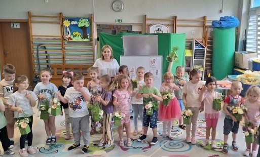dzieci wraz z panią pozują do zdjęcia grupowego w tle wystrój sali gimnastycznej oraz tablica pokazowa zajęć