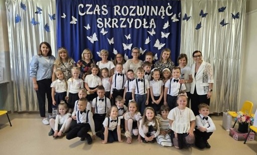 grupa odświętnie ubranych dzieci pozuje do zdjęcia grupowego wrz z paniami w tle dekoracja na zakończenie roku