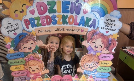 Dwie dziewczynki pozują za kolorową planszą z napisem Dzień Przedszkolaka i ilustracjami dzieci, tęczy oraz słońca.